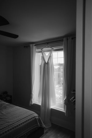 A white wedding dress hangs in a window, captured in black and white photography.