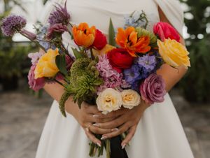 A vibrant bridal bouquet featuring colorful ranunculus, roses, and wildflowers against a white dress background.