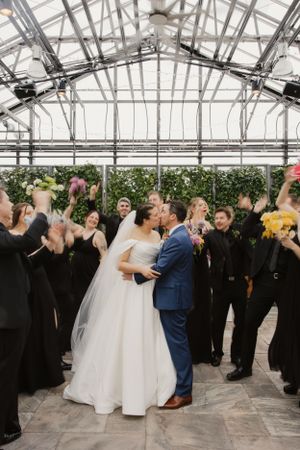 Wedding celebration in a greenhouse venue with guests cheering as newlyweds kiss under the glass ceiling and greenery.