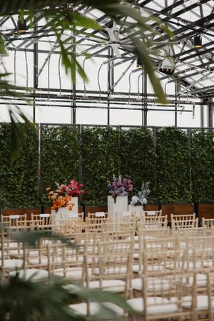 Indoor greenhouse wedding venue with white chairs and colorful floral arrangements against a wall of green foliage.