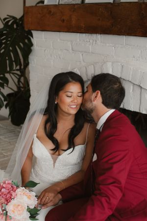 Romantic wedding moment with bride in white dress and groom in burgundy suit near white brick fireplace.
