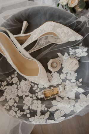 Elegant white lace wedding shoes with veil and floral decorations on grey background.