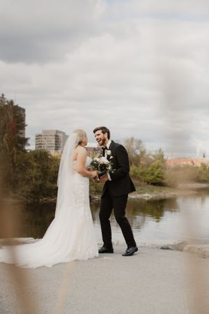 Newlywed couple sharing romantic moment by lake with bride holding white bouquet.