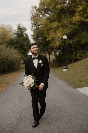 Groom in classic black tuxedo holding bouquet while walking on park path.