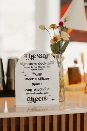 Bar menu sign displaying cocktail options with fresh flowers in a vase.