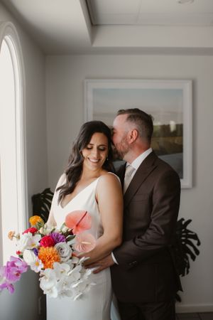 Intimate moment between newlyweds with vibrant bouquet against white interior.