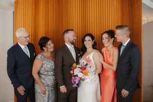 Wedding party group photo against orange wall with colorful bouquet.