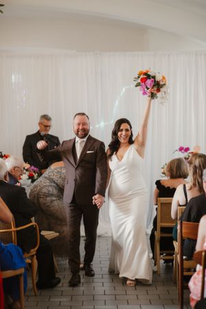 Wedding couple celebrating down aisle with raised bouquet and happy guests.