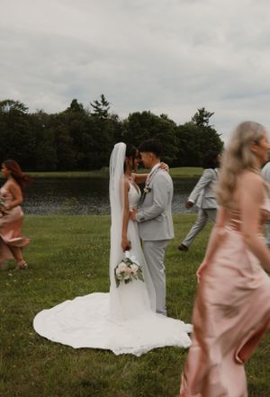 A couple shares their first kiss as newlyweds by a lake while wedding party moves around them.