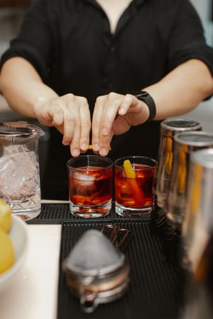 A bartender prepares cocktails with red liquor and citrus garnishes on a black bar mat.