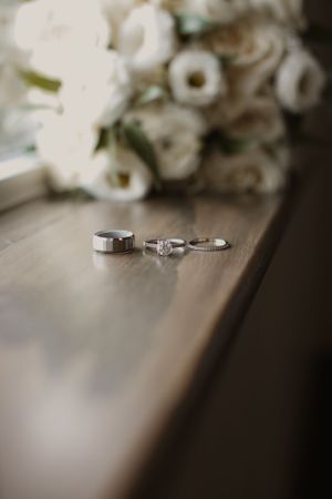 Three wedding rings arranged on a wooden surface with blurred white roses in the background.