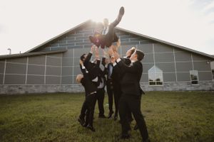 Groomsmen in dark suits toss someone in the air outside a building at sunset.