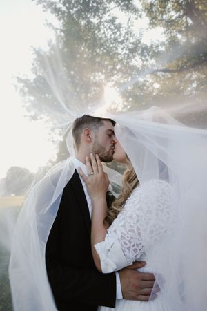 Couple embracing under flowing veil with sunlight streaming through trees.