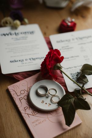 Wedding rings displayed on a small plate with a red rose and invitation cards on a wooden surface.