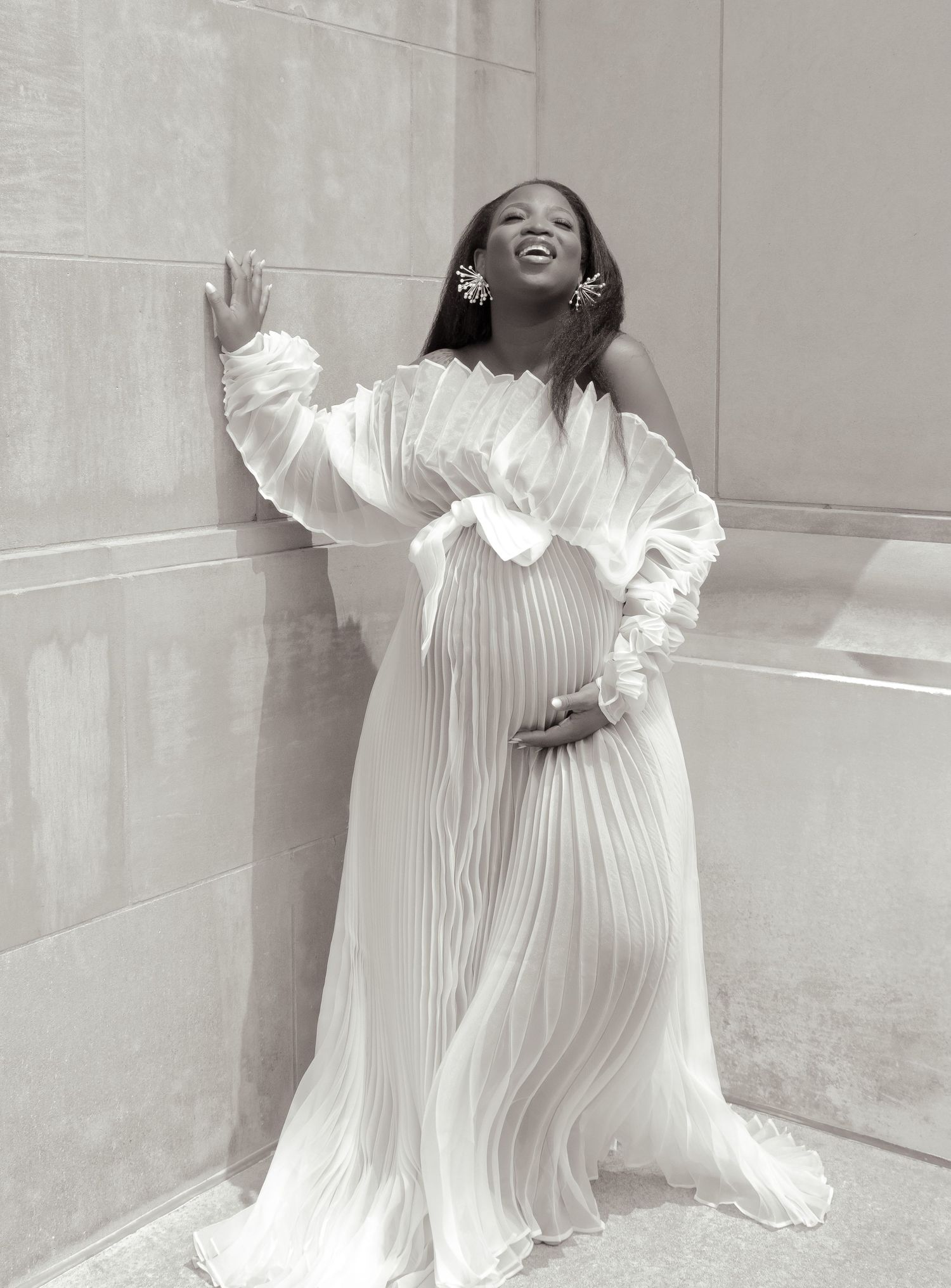 A joyful person in a flowing white pleated maternity gown laughs while posing against a light colored wall.