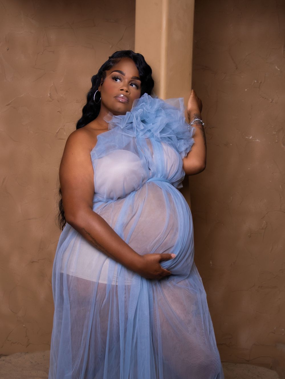 Maternity photo showing ethereal light blue tulle gown against earth-toned wall.