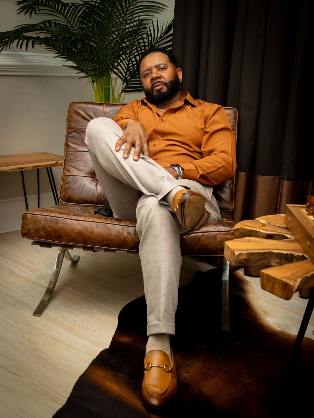 Stylish individual relaxing in brown leather lounge chair wearing orange shirt, light pants and brown loafers.