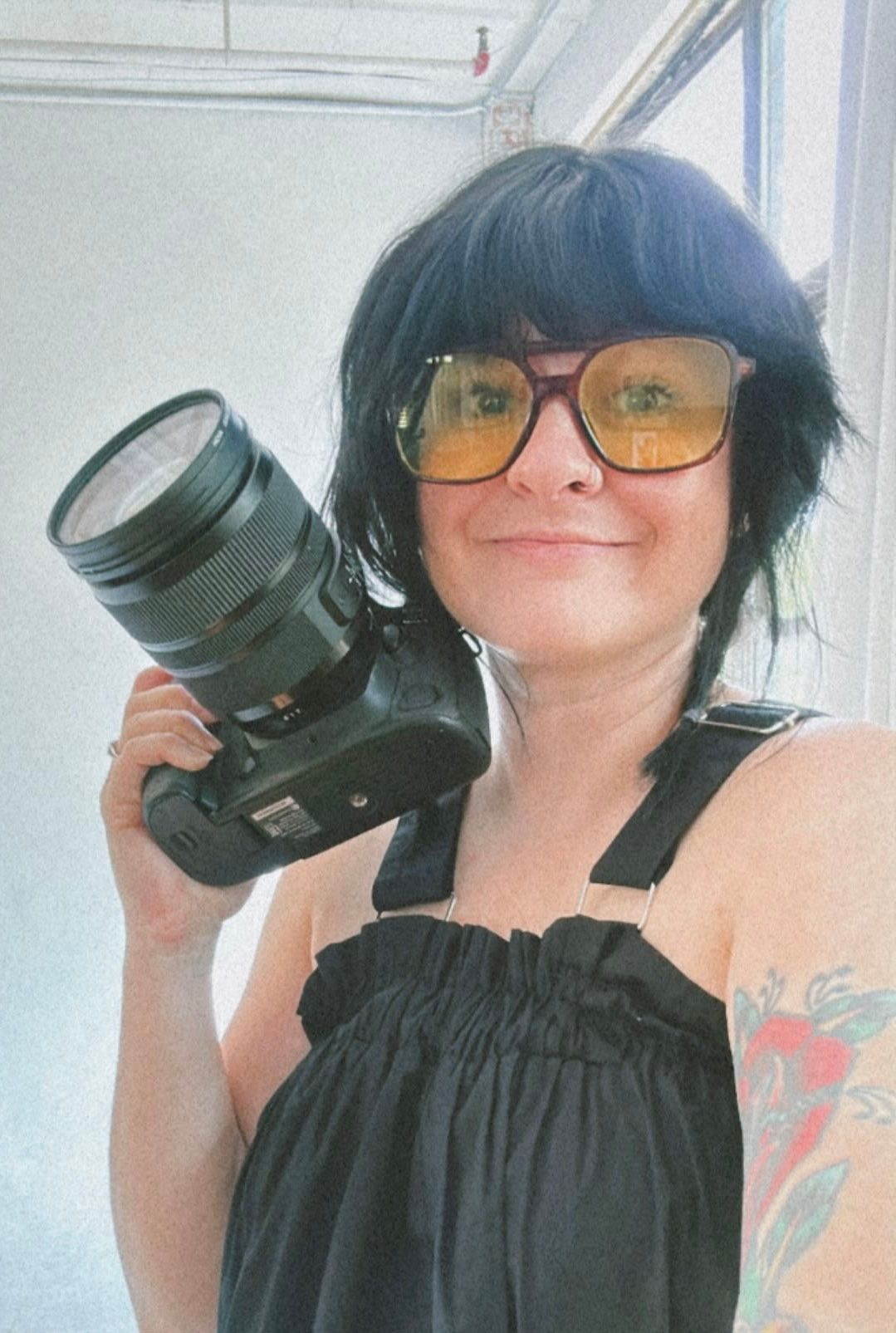 Woman with black hair and yellow sunglasses holds a DSLR camera, wearing a black top with a tattoo visible.