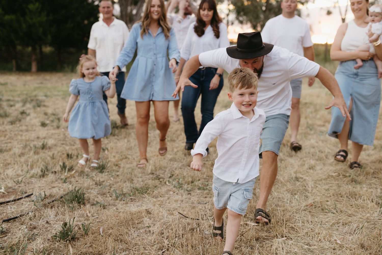 Family group photo outdoors with father in black hat chasing laughing boy across dry grass field at golden hour.