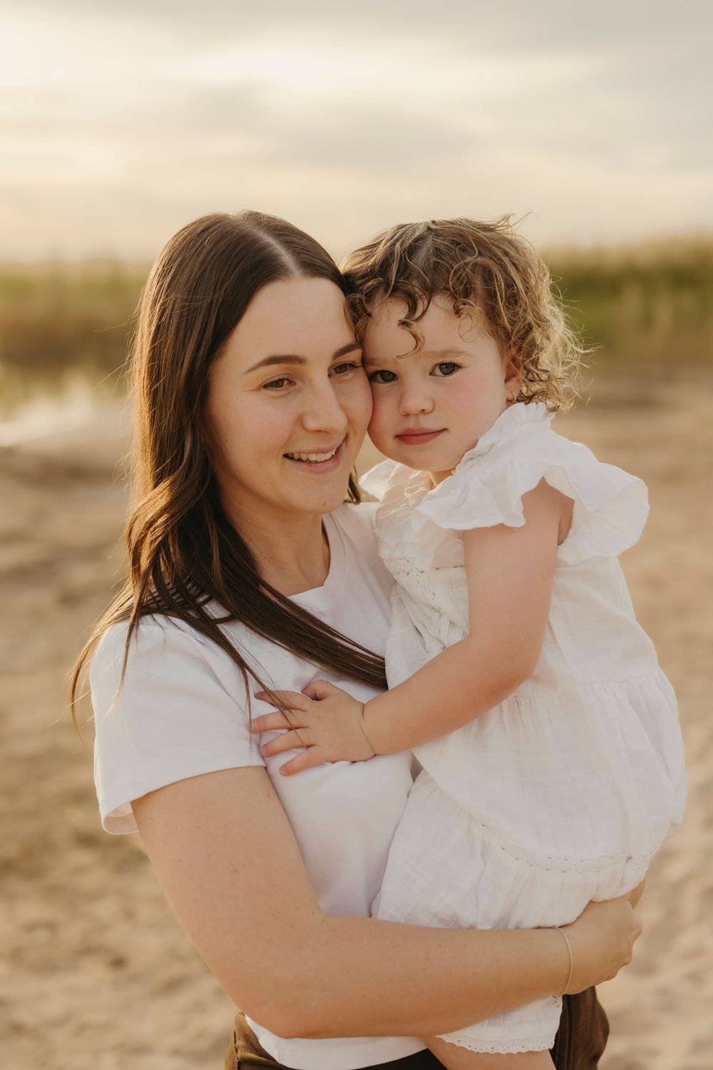 A young mother smiles warmly while holding her curly-haired toddler daughter at sunset on a beach.