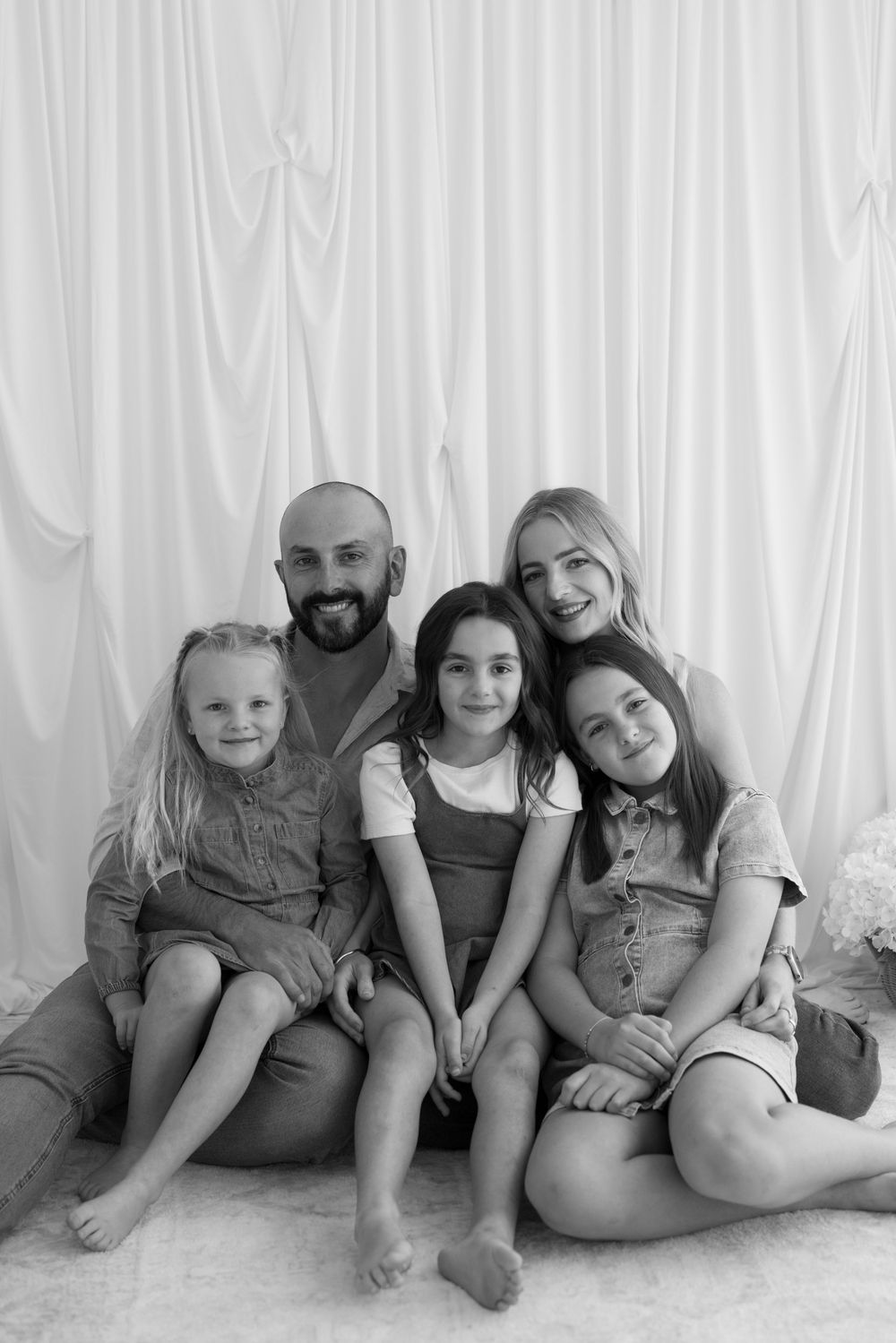 Black and white family portrait of parents with three daughters sitting together against white draped backdrop.