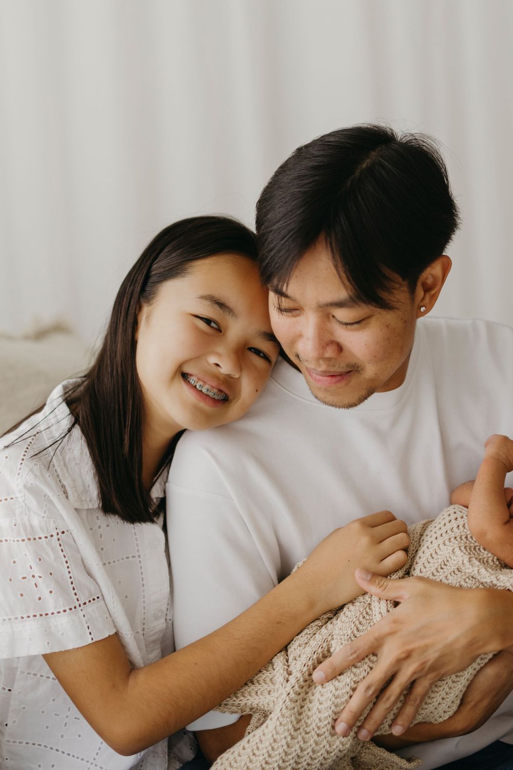 Smiling Asian couple holding newborn baby together in soft natural light.