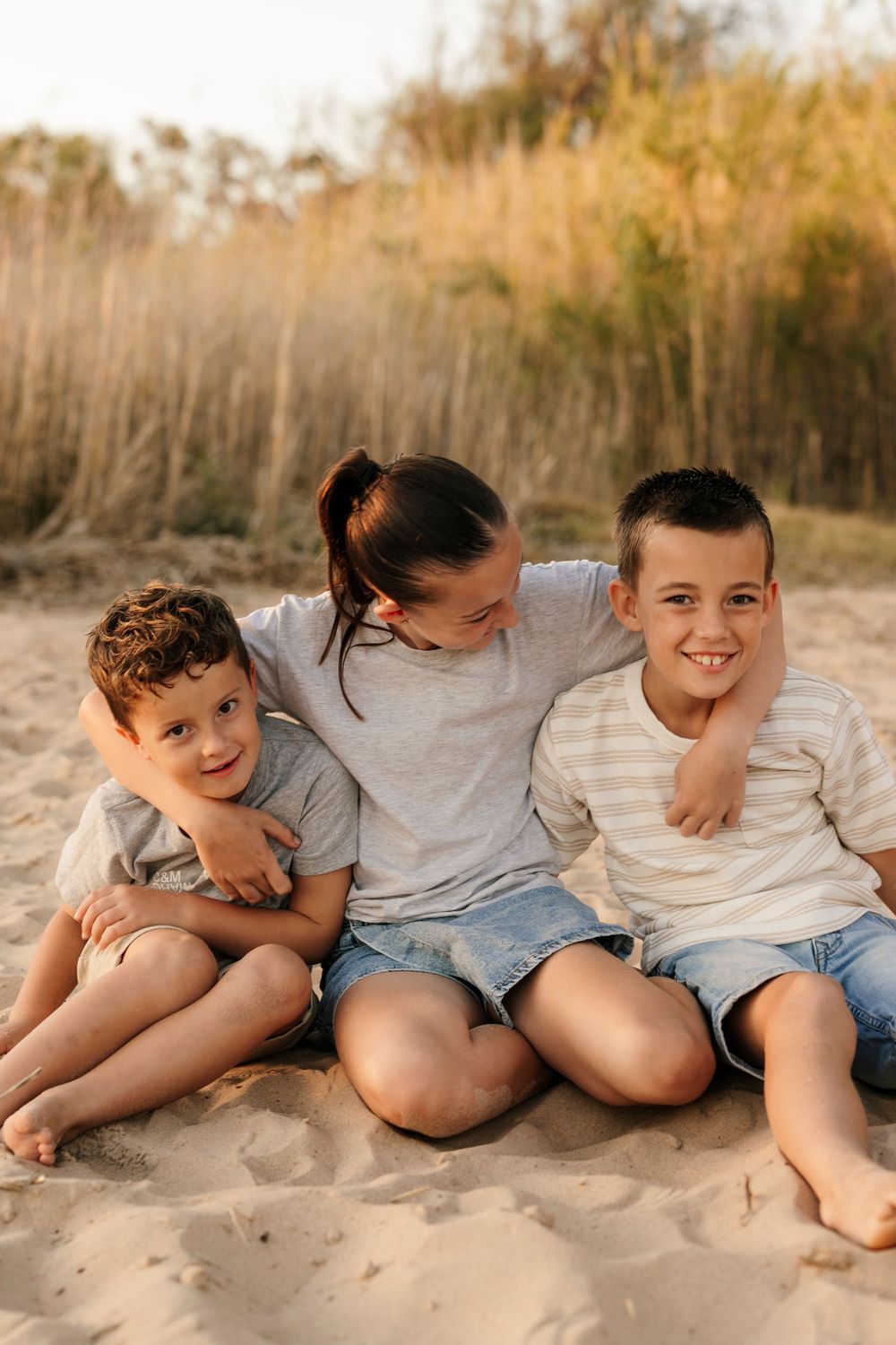 Three siblings sitting together on sandy beach at golden hour, smiling at camera.