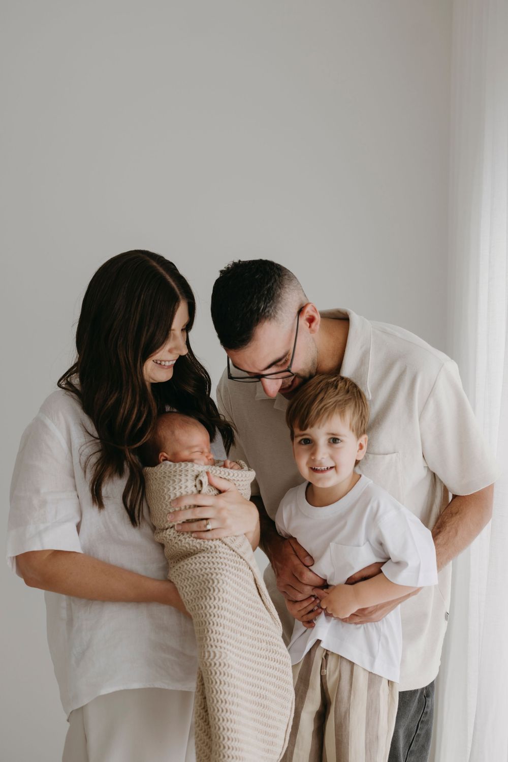 A family of four gathers lovingly around their swaddled newborn in a bright white studio.