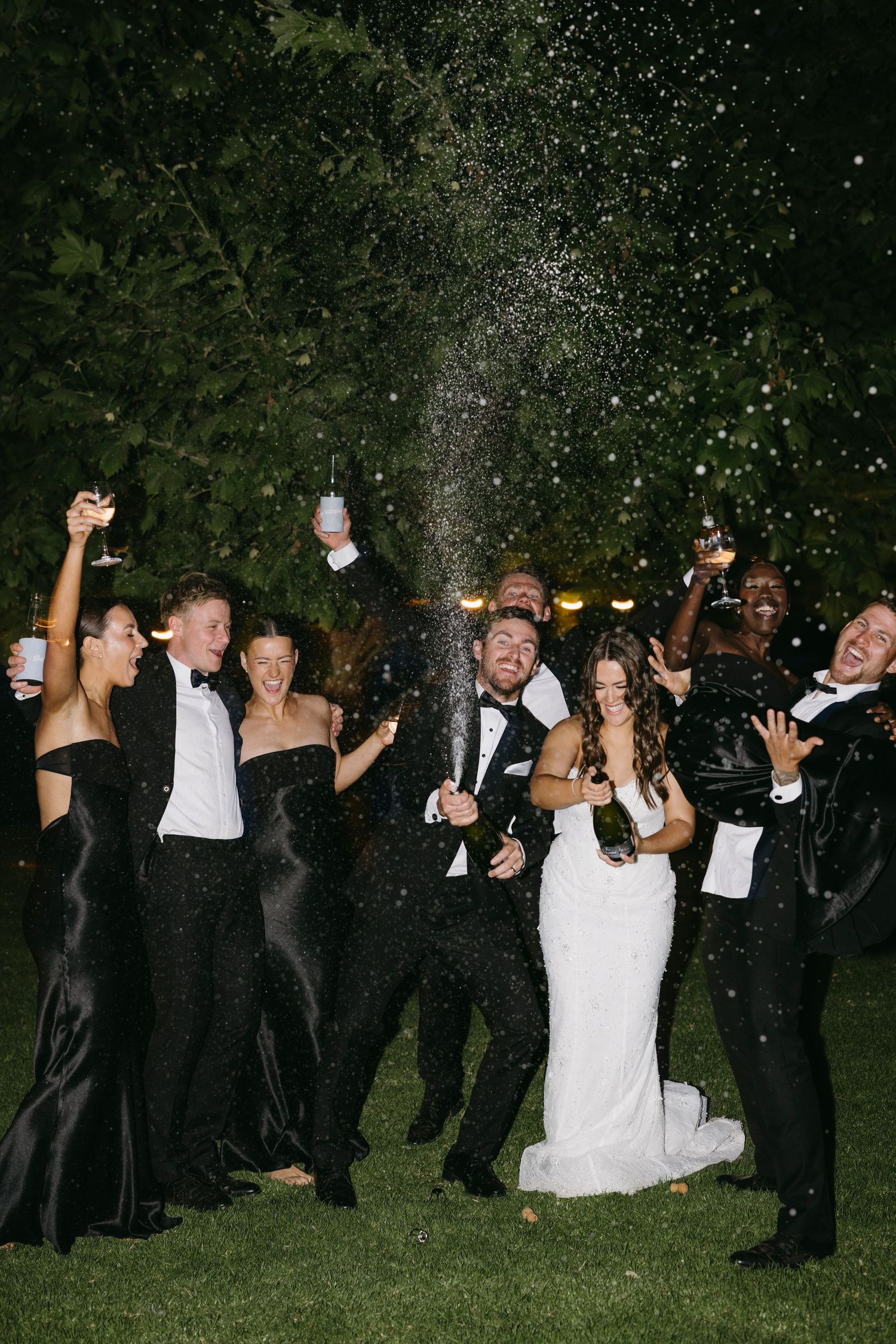 Wedding party celebrating at night with champagne bottle spraying as bride and groom laugh with guests outdoors.