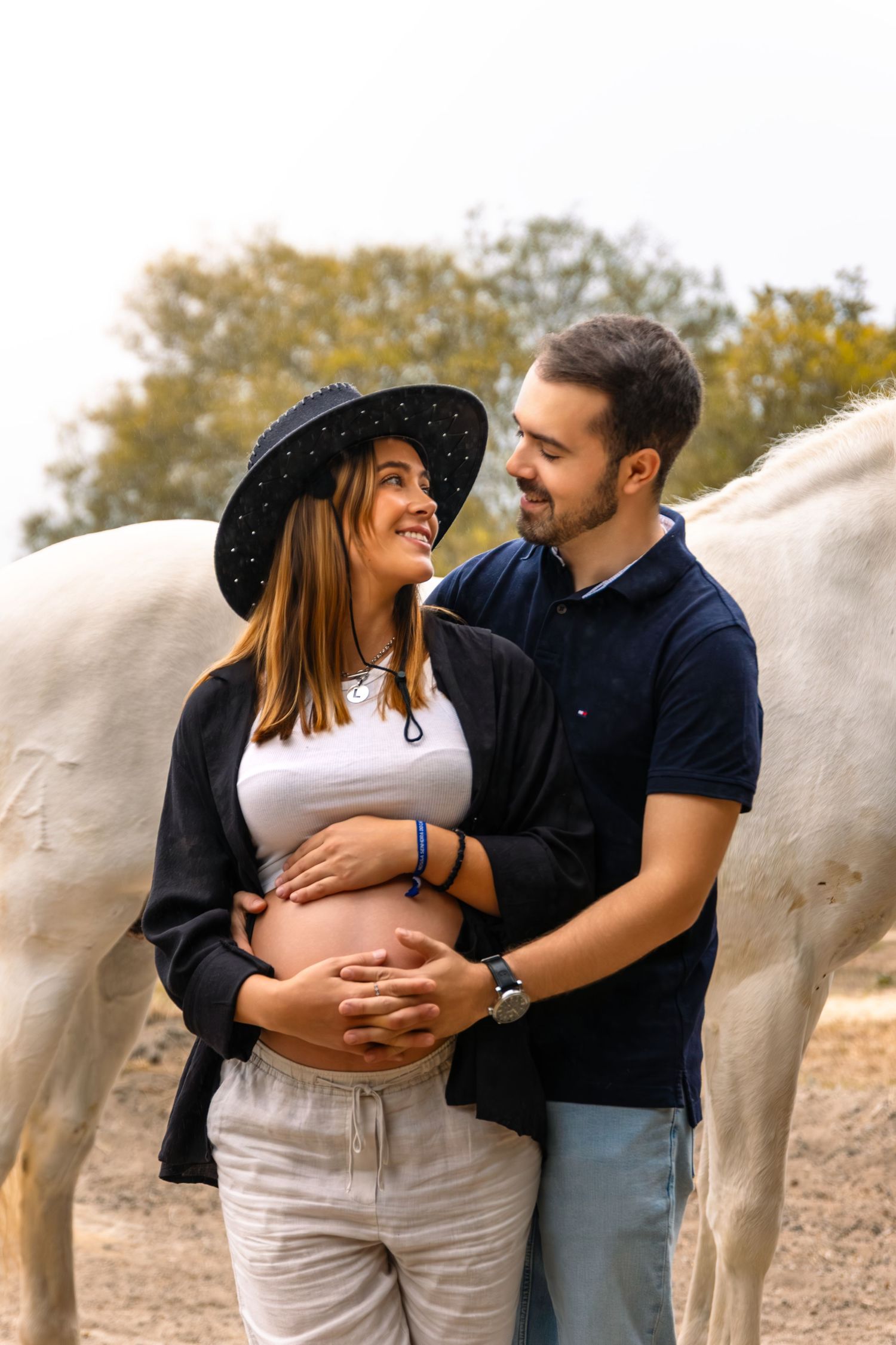 Um casal partilha um momento íntimo num campo com cavalos brancos ao fundo.