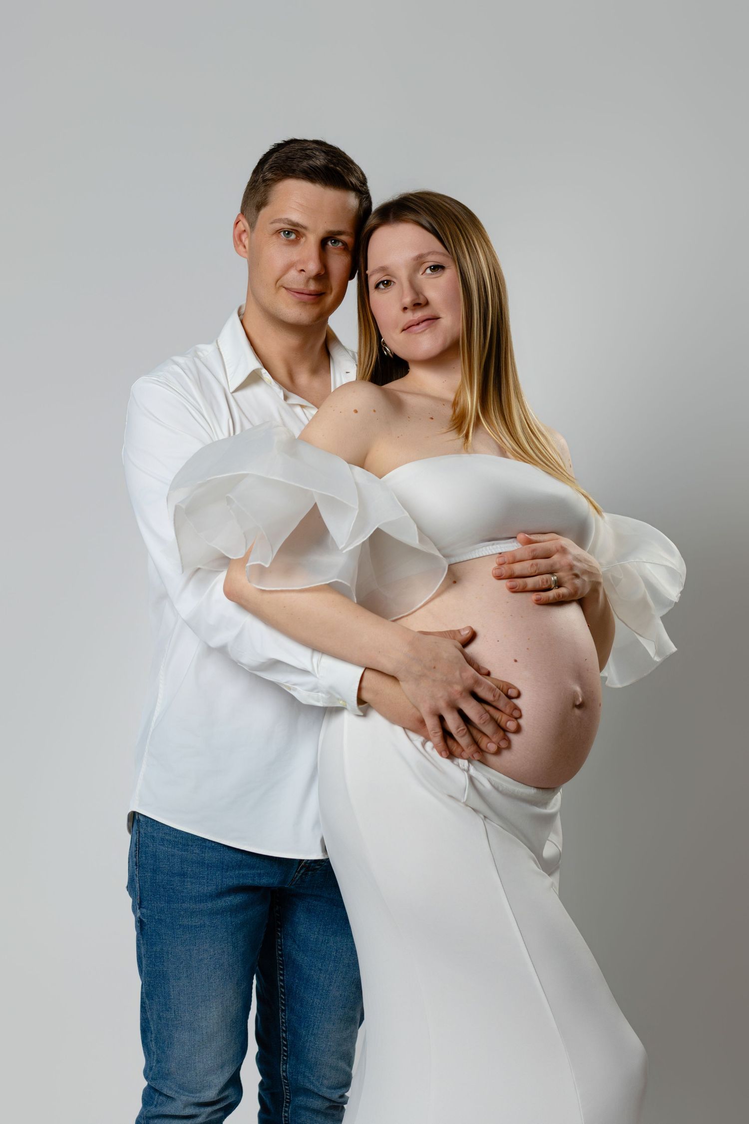 A couple in white clothing poses together for a maternity photo shoot against a light gray background.