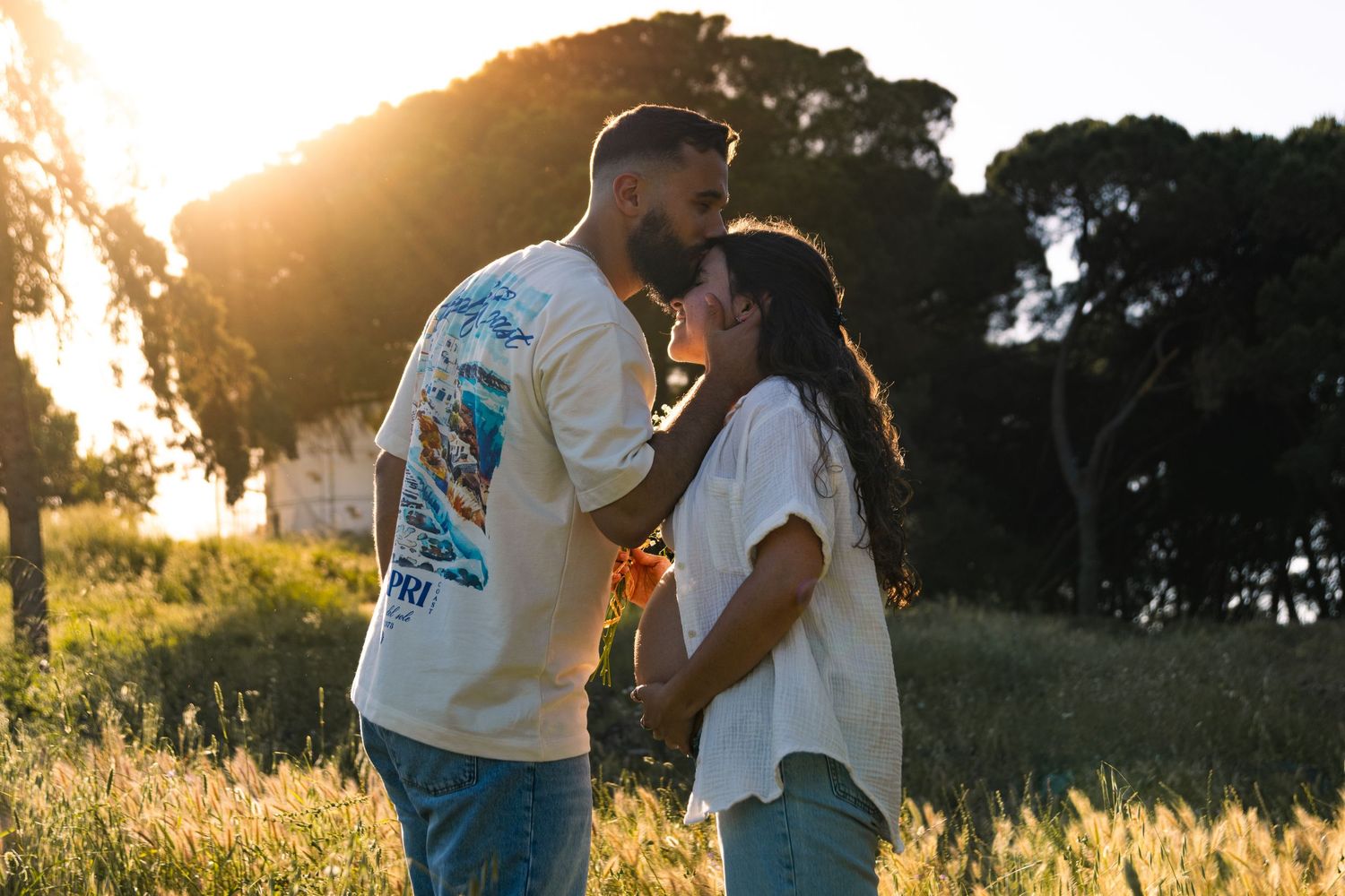 Couple embracing in golden sunlit field at sunset with trees in background.