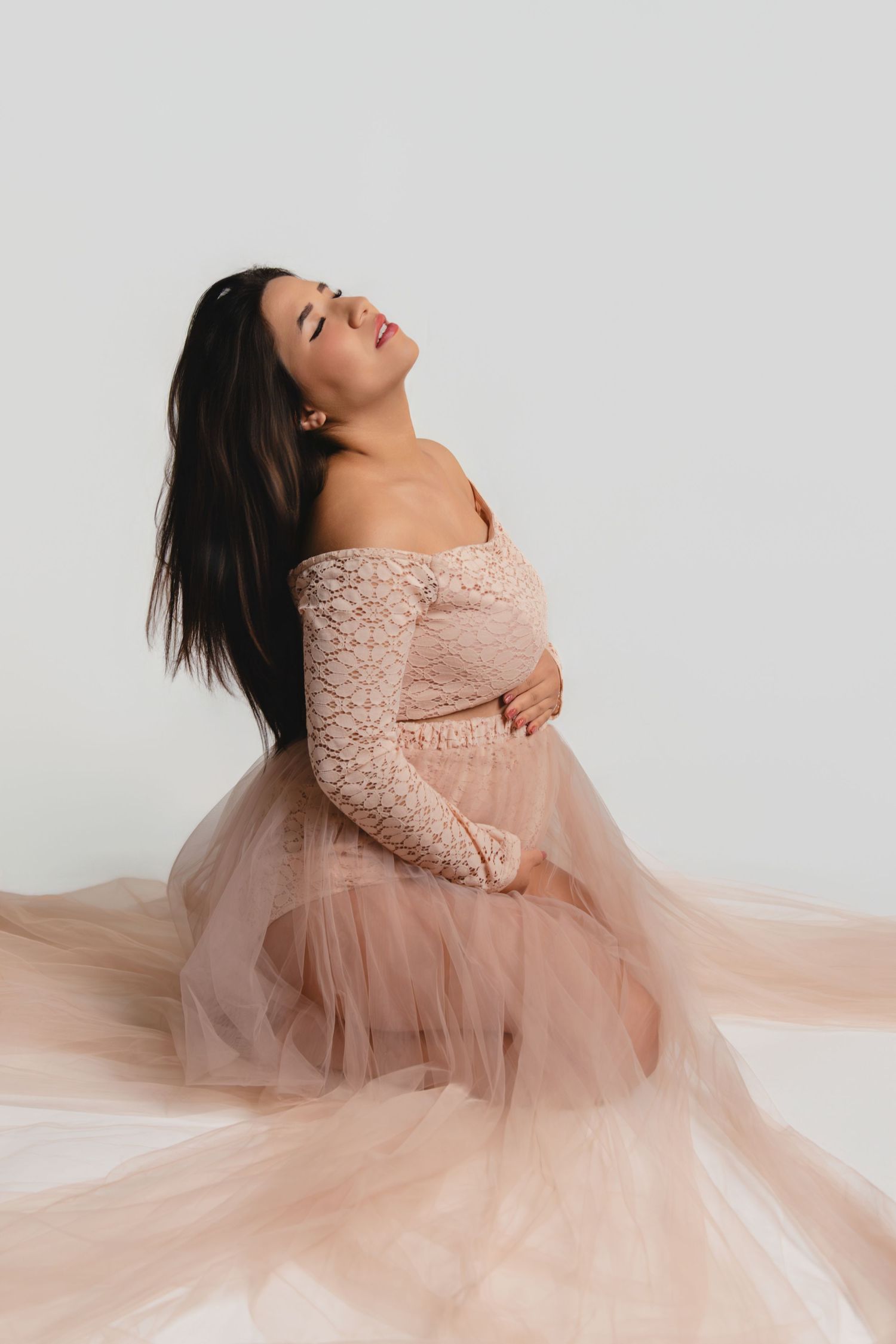 Maternity portrait in flowing pink lace dress against white background.