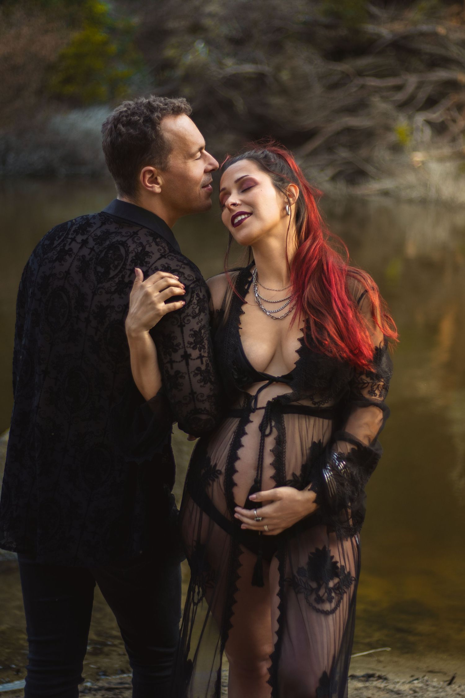 Couple in black gothic attire share an intimate maternity moment by a lake.