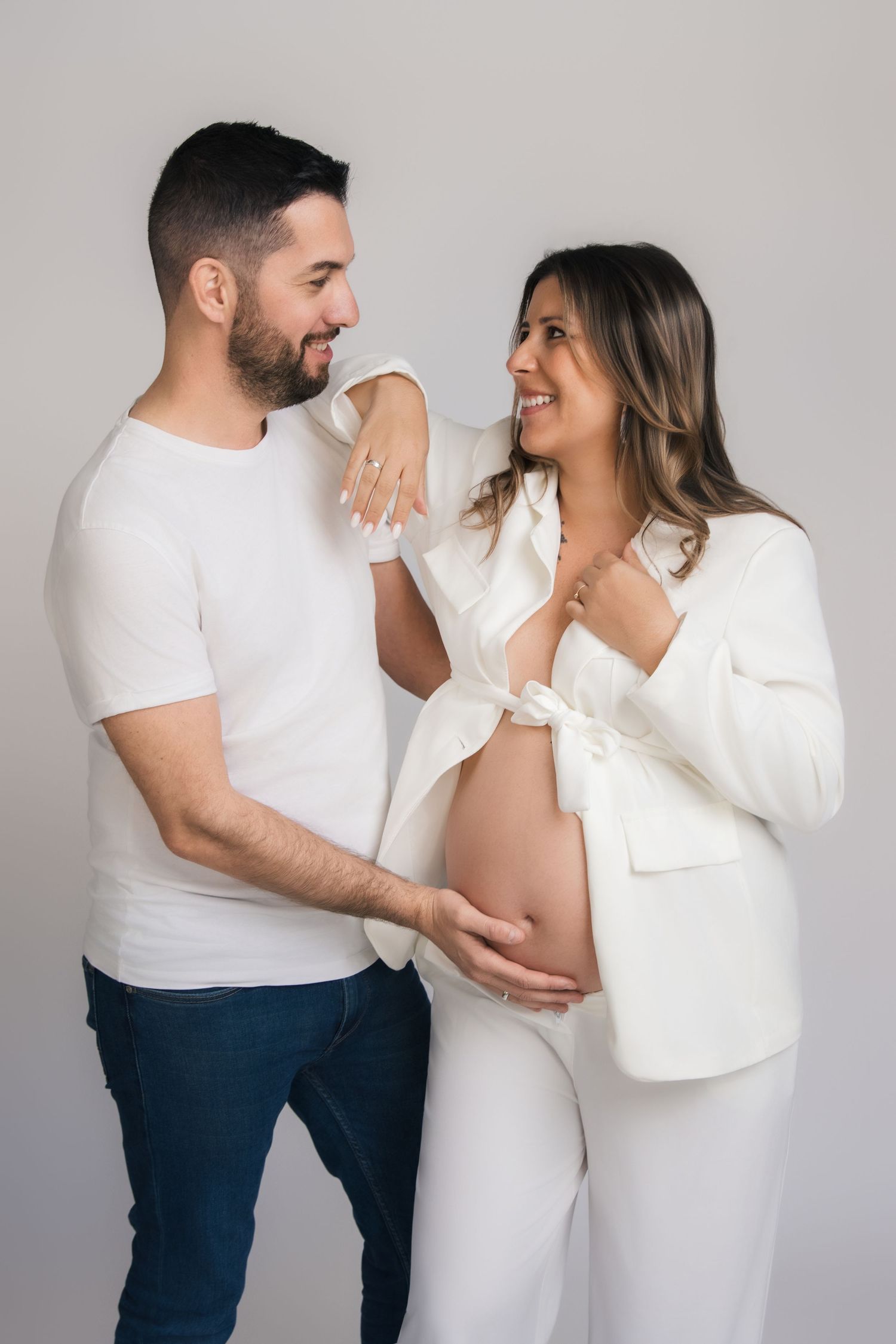 A couple in white outfits and jeans share playful candid moments against a gray backdrop in a photo series.