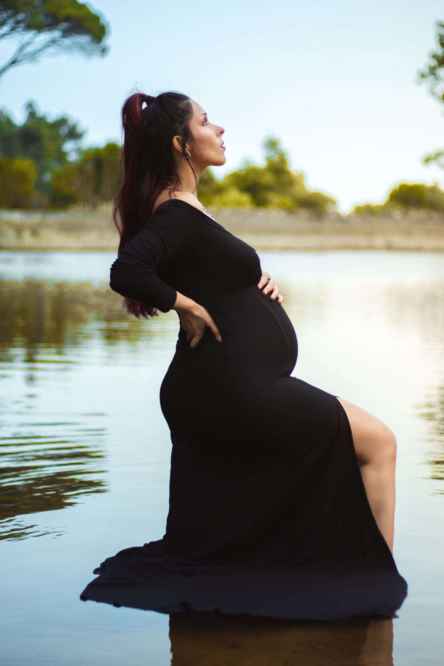 Silhouette of pregnant woman in black dress poses elegantly by water at sunset.