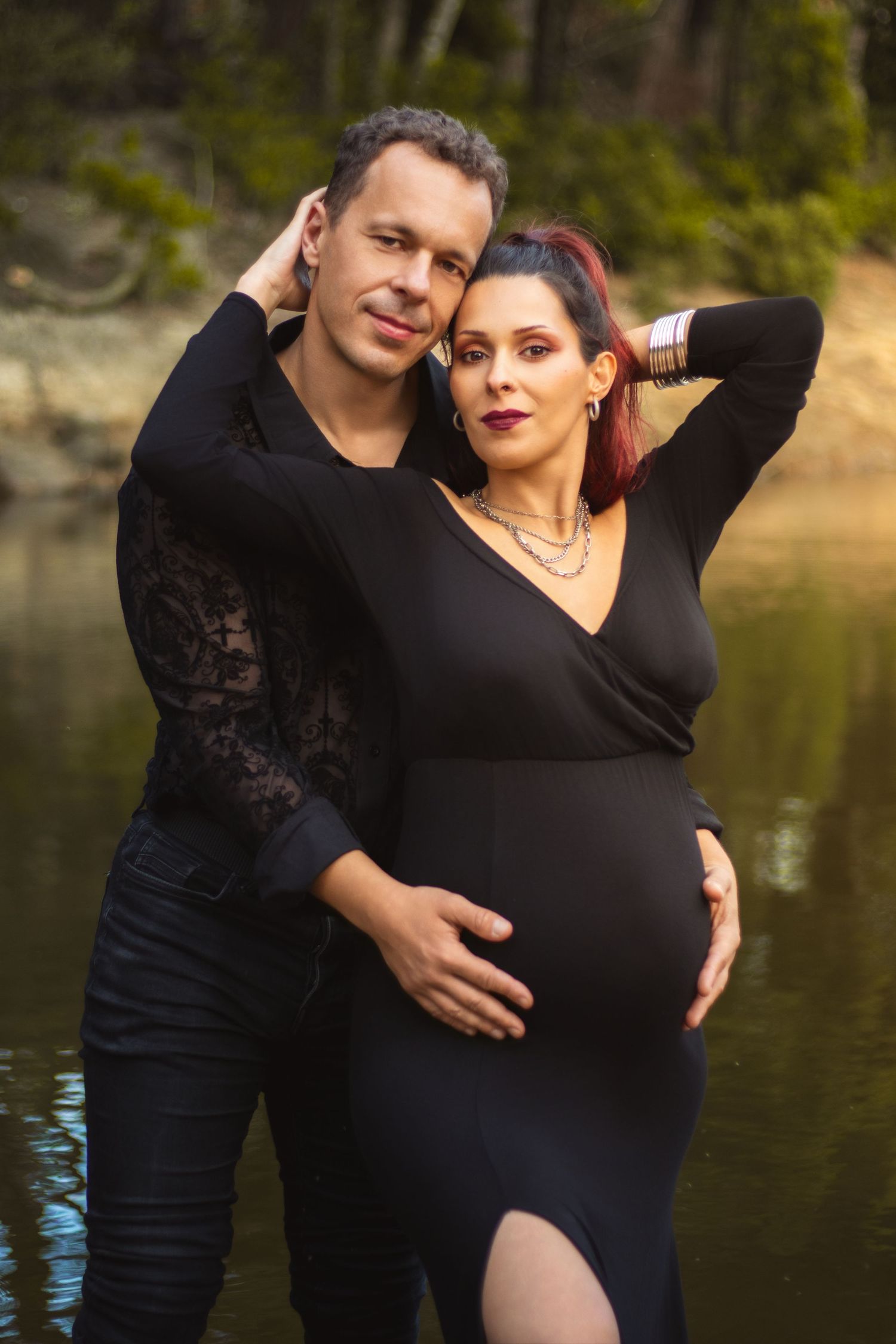 Maternity photoshoot by a lake featuring a couple in black outfits embracing against a natural outdoor backdrop.