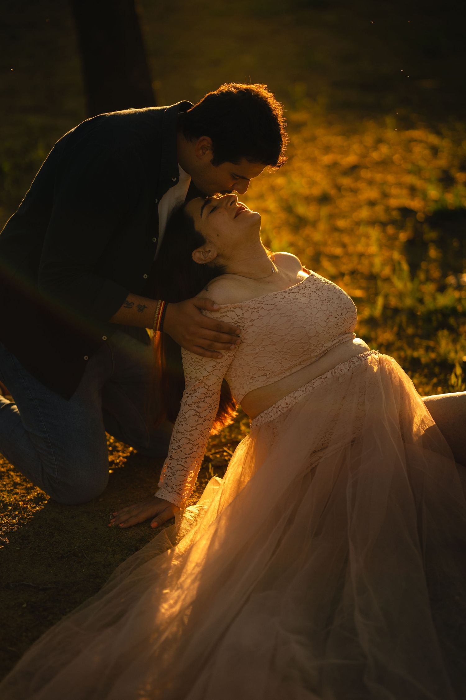 Bride in white lace gown sits on ground as groom leans in for a kiss during golden hour outdoor wedding.
