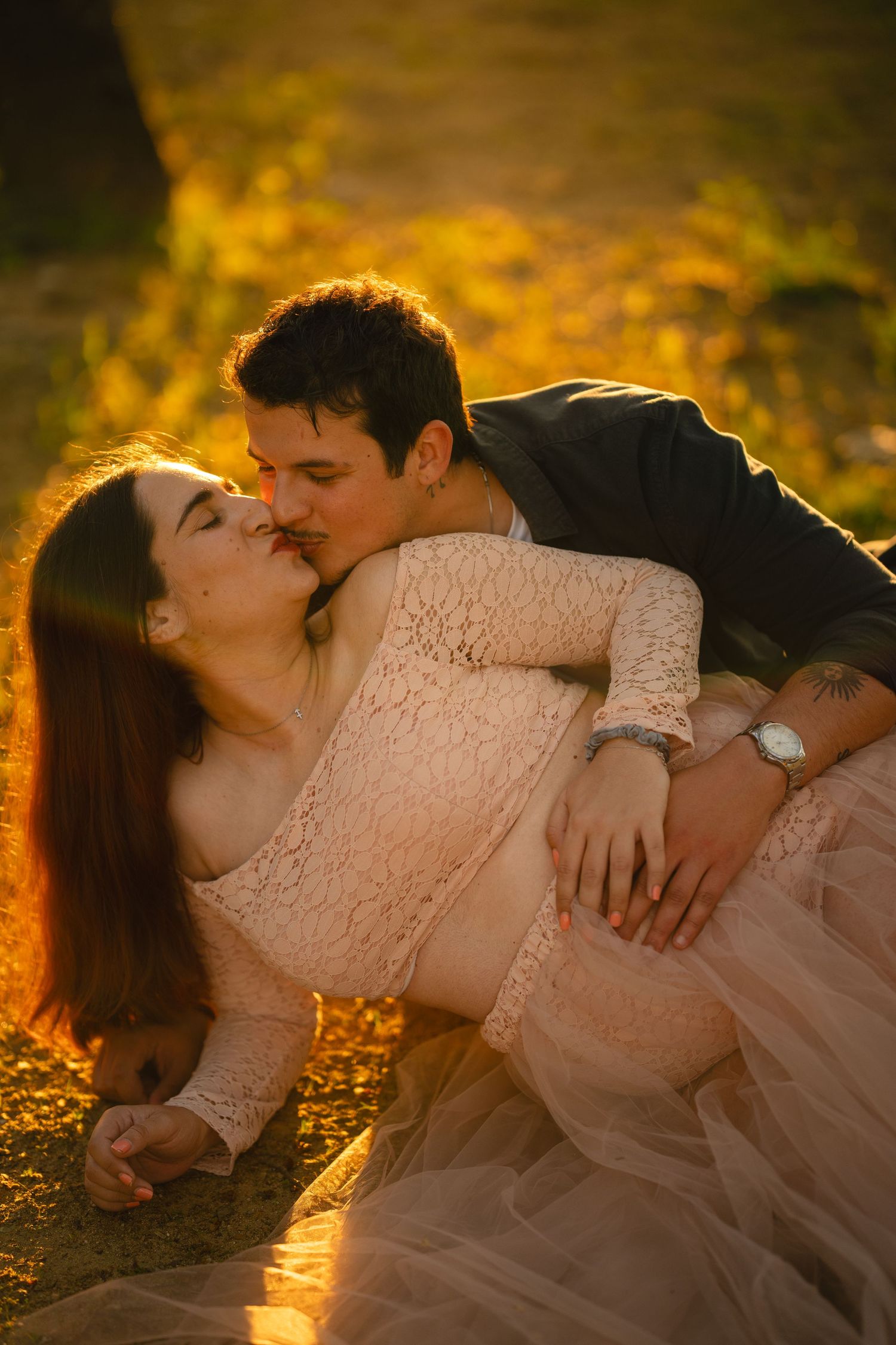 Couple sharing a romantic kiss outdoors during golden hour, woman in lace dress, man in dark jacket, autumn setting.