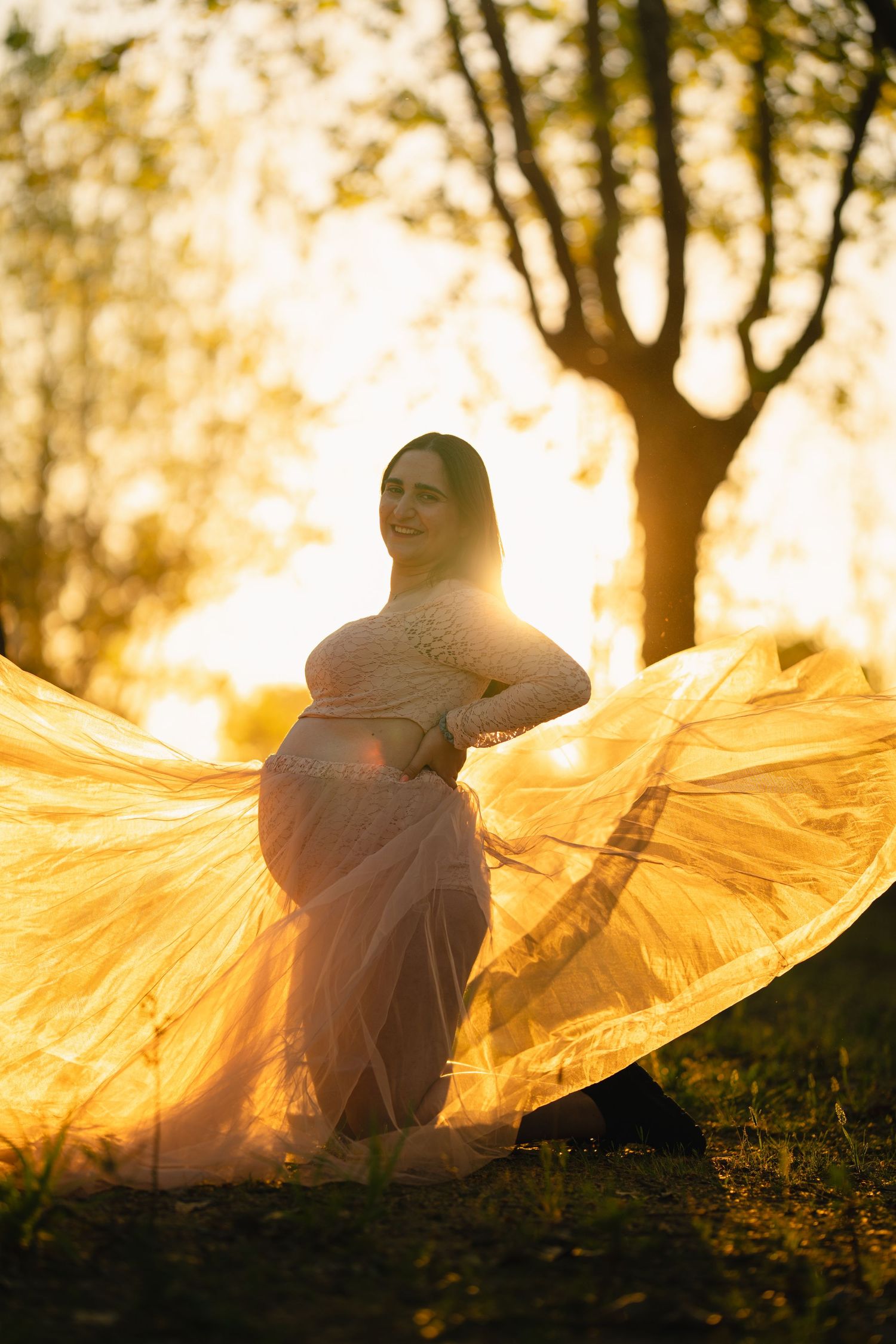 Pregnant woman in flowing golden dress poses outdoors at sunset with fabric billowing in warm backlit glow.