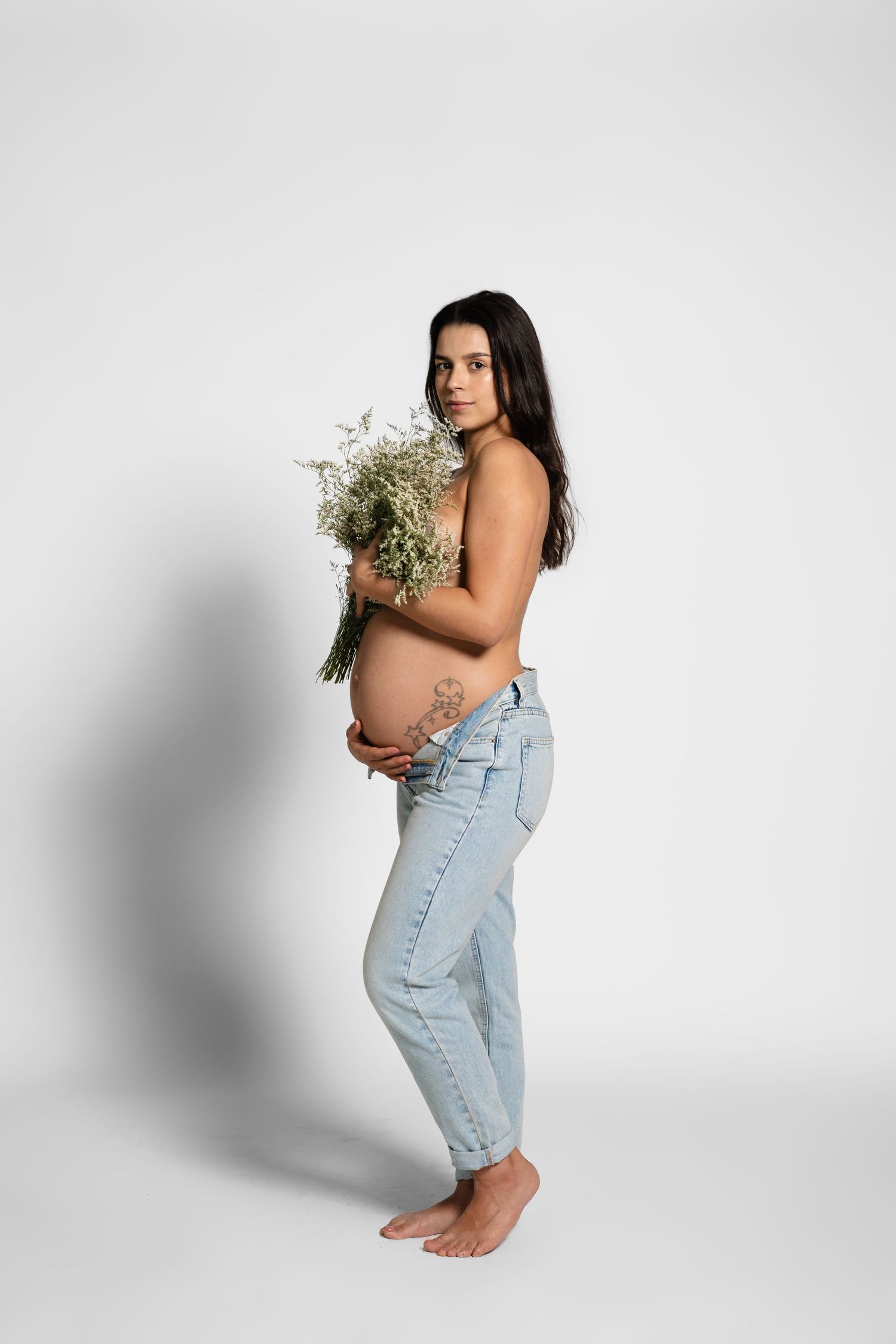 Side view of pregnant woman in denim holding dried flowers against white background.