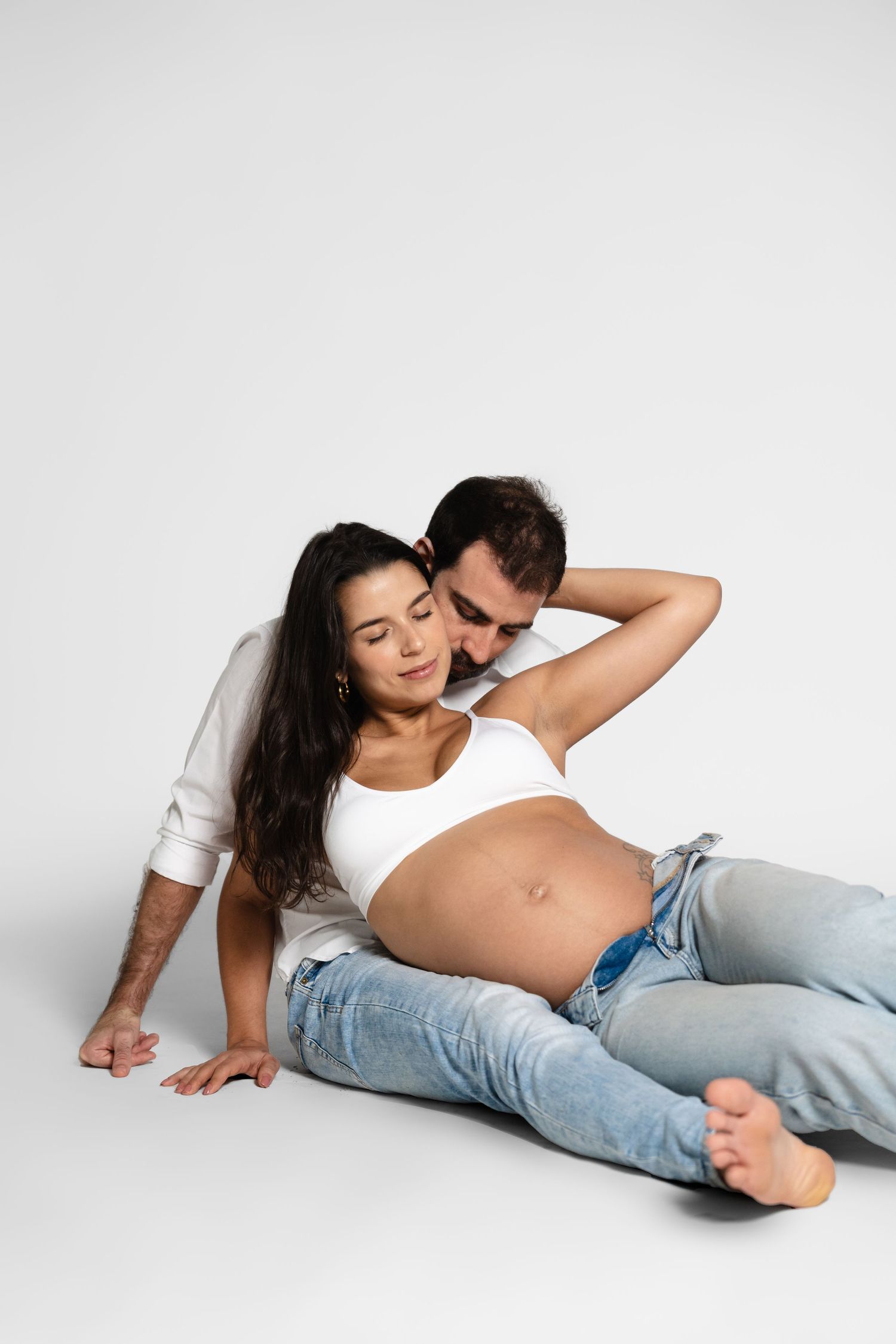 Artistic maternity photoshoot with couple resting on white studio floor.