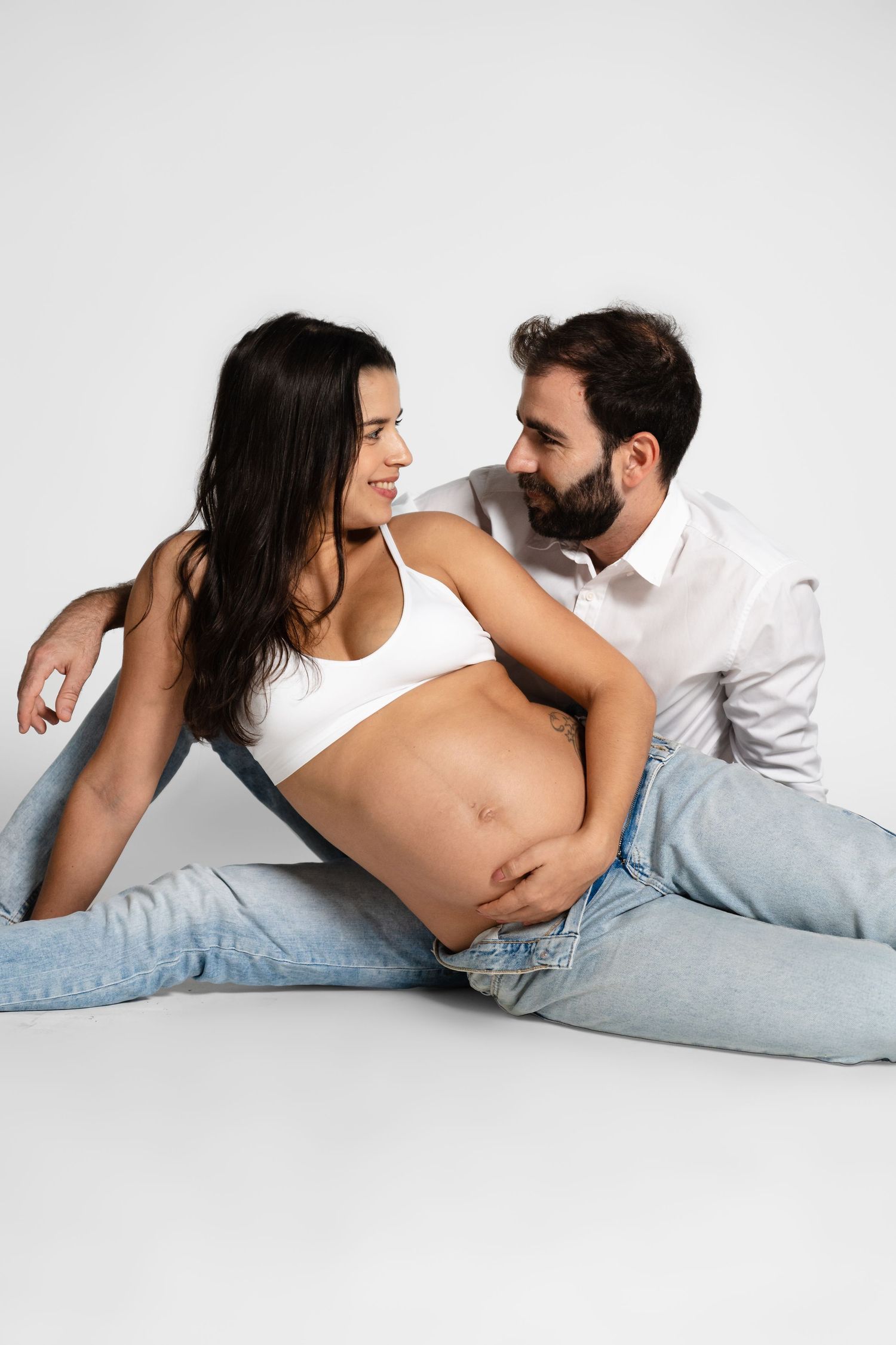 Intimate maternity photo series showing a couple lying down in casual attire against white background.