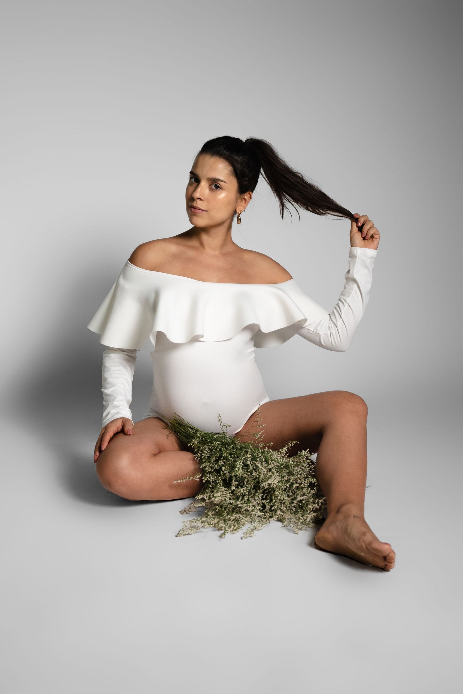 Maternity photoshoot featuring woman in white off-shoulder dress with greenery.