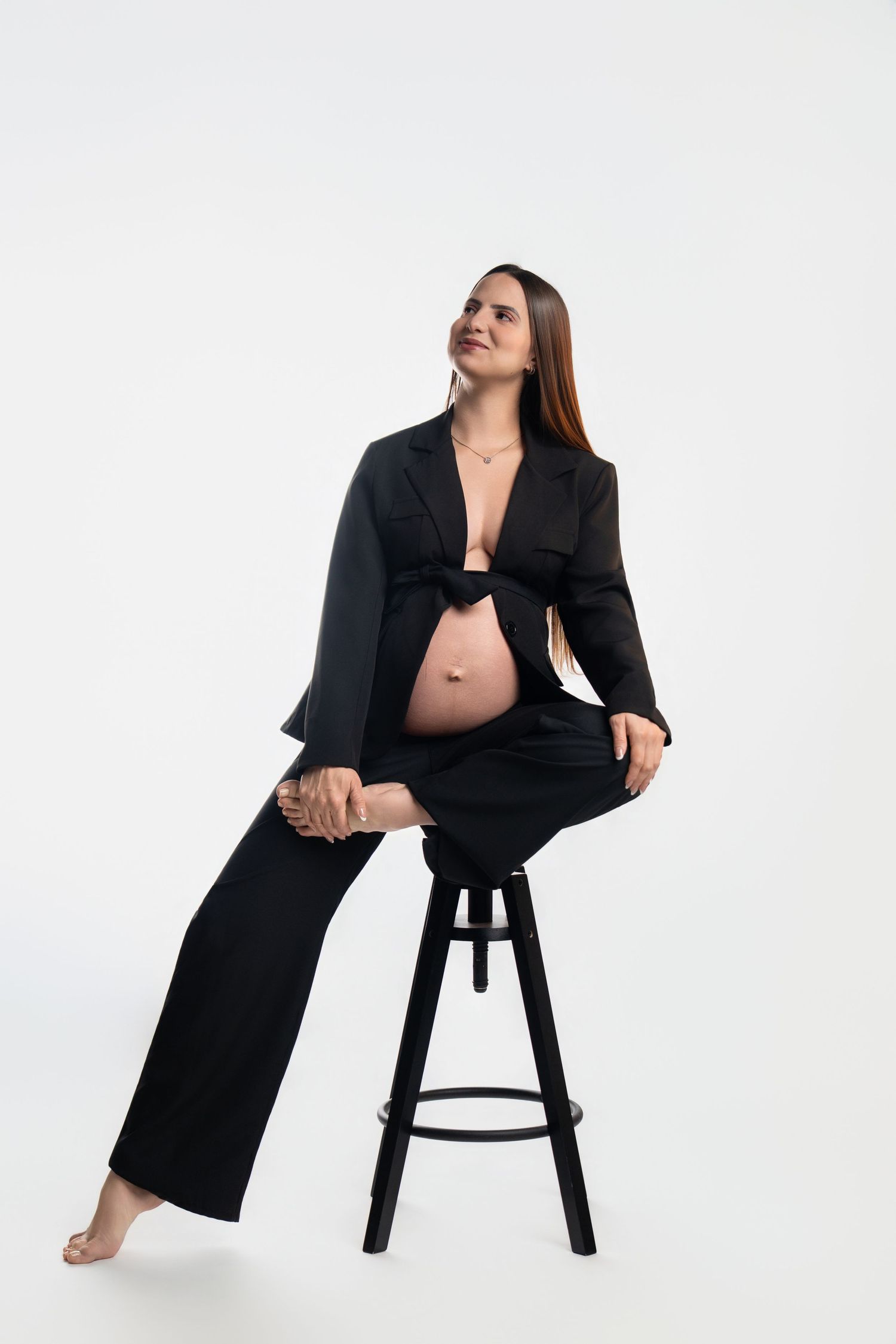 Minimalist maternity portraits with black blazer and stool against white backdrop.