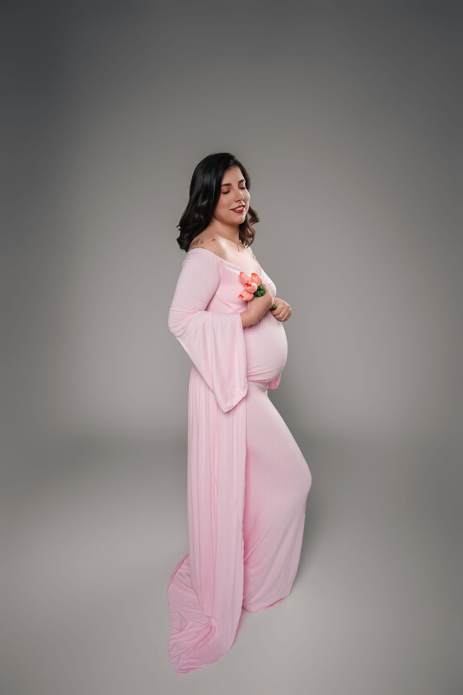 Smiling pregnant woman in elegant pink maternity gown holding pink flowers, studio portrait on gray background.