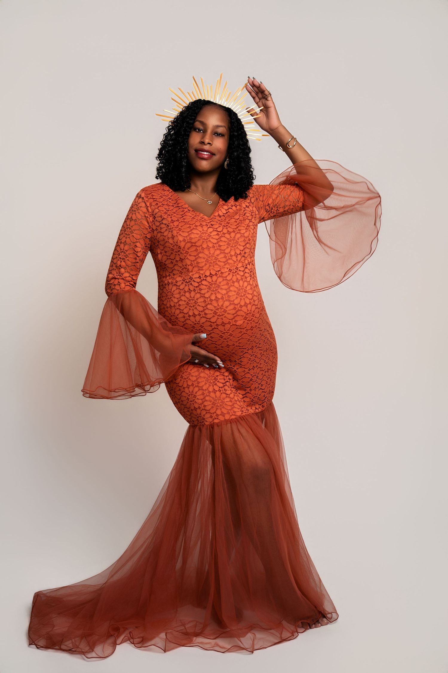 Elegant maternity photo of woman in flowing rust-colored lace gown with sheer bell sleeves.