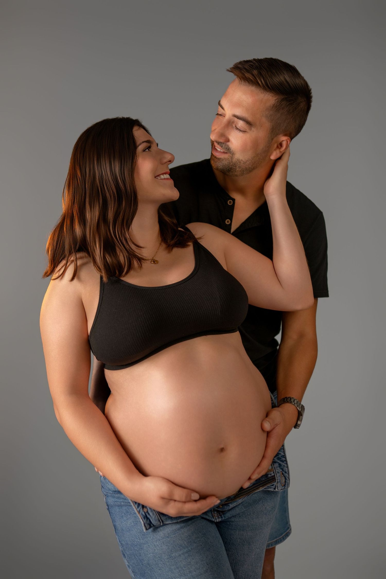 Intimate studio maternity session with couple in casual attire against grey background.
