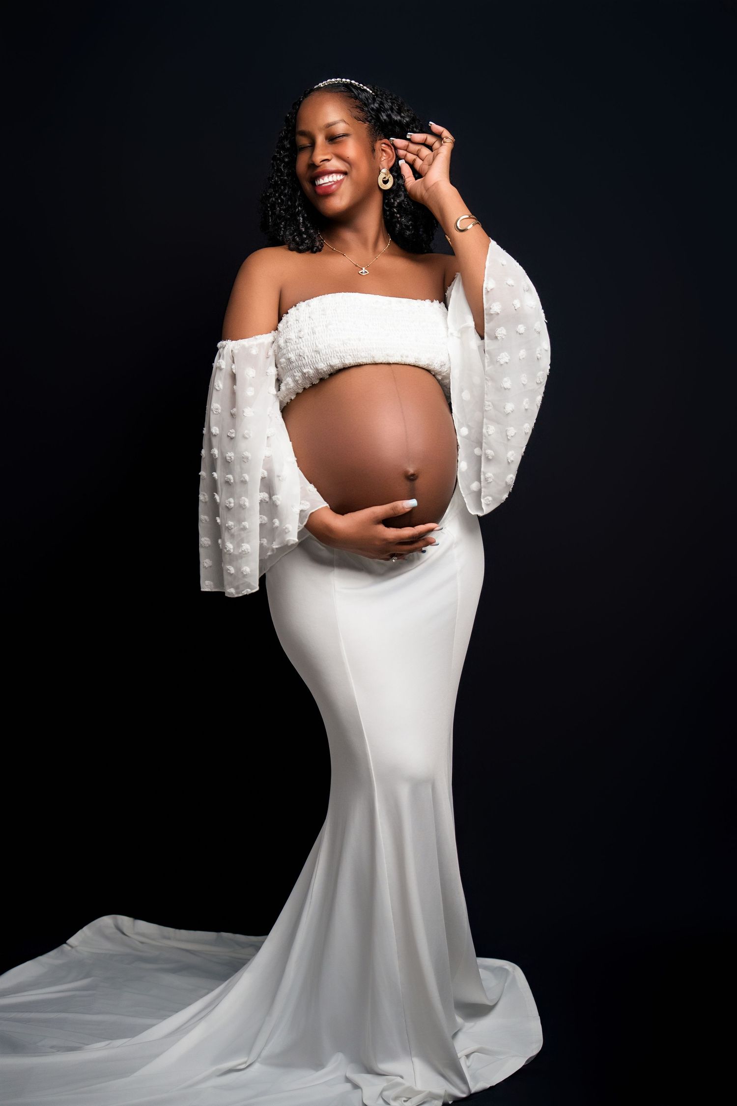Stunning maternity portrait in white off-shoulder gown with bell sleeves against black backdrop.
