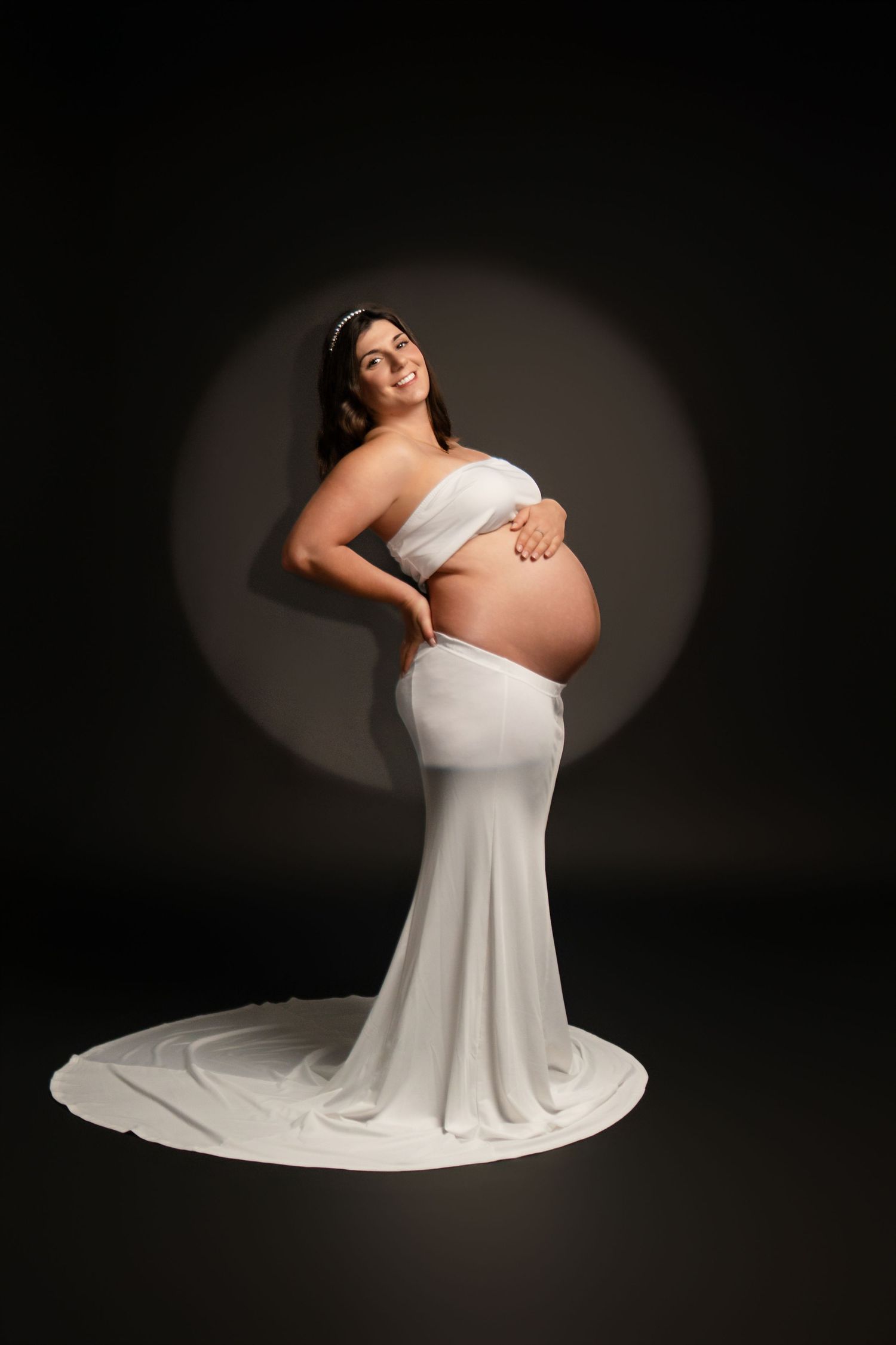 Elegant maternity portraits in flowing white dress against dark studio backdrop.